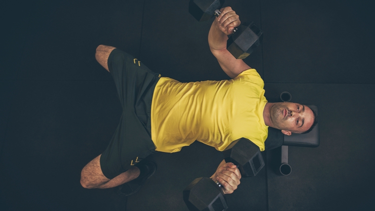 man on flat bench in gym stretching chest with dumbbells