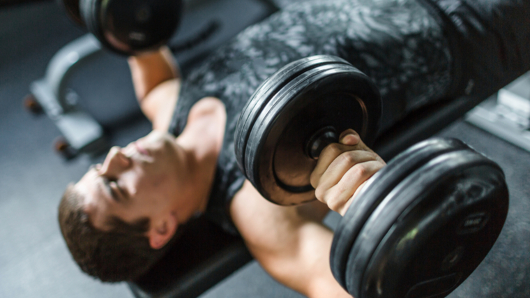 man in gym lying on bench lifting heavy dumbbells