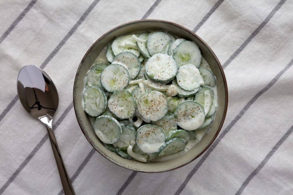 A bowl of sliced cucumbers mixed with creamy dressing and sprinkled with pepper sits on a cloth with gray stripes. A spoon is placed next to the bowl. MyFitnessPal Blog