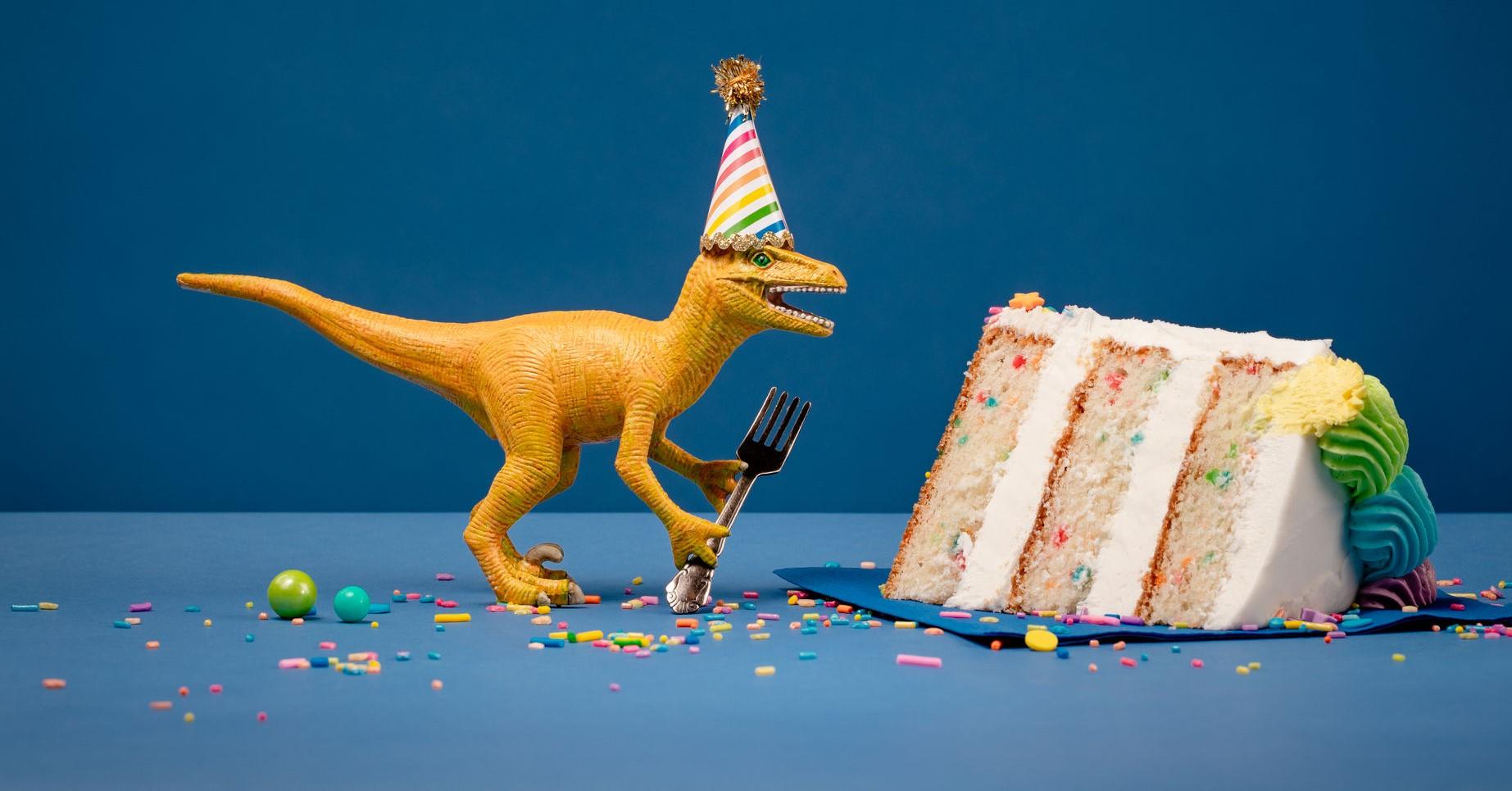 Toy Dinosaur holding a fork next to a slice of birthday cake on a blue background.