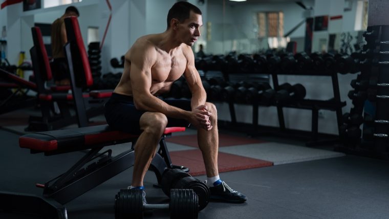 Man sitting on a weight bench with a pair of dumbbells near his feet.